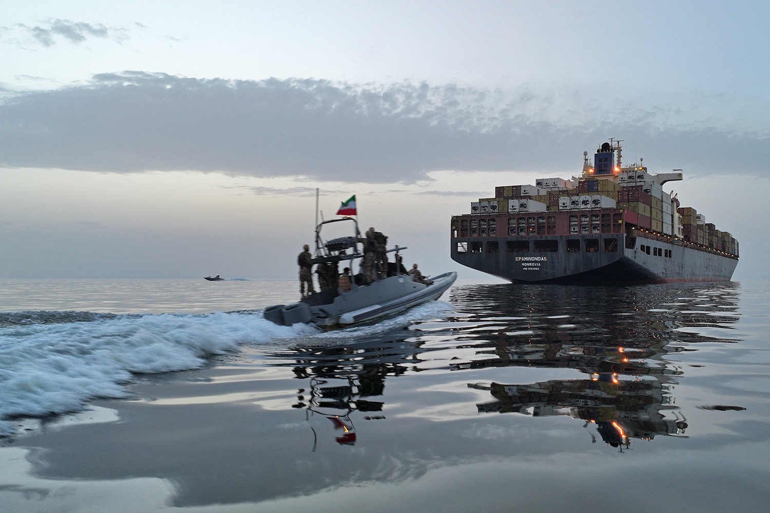 A small, dark motorboat carrying several people in military gear speeds across calm water toward a massive container ship. The container ship is loaded with colorful cargo crates and occupies a large portion of the right side of the frame. The sky is pale during dusk or dawn, reflecting on the water’s surface.