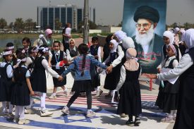 About two dozen schoolchildren around age 8 or so in black-and-white school uniforms play around on a concrete square with the U.S. and Israeli flags painted on the ground. Many of the children hold hands as they spin in a circle. Nearby, a banner with a portrait of Khamenei watches over the children.