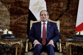 Sisi sits in a golden chair in front of the Egyptian flag, wearing a dark suit and dark red tie. His hands are resting on his thighs.