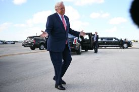 Trump stands on a tarmac with his arms out wide and eyes closed.