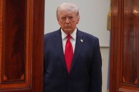 Donald Trump is seen wearing a blue suit and a red tie, standing in a doorway.